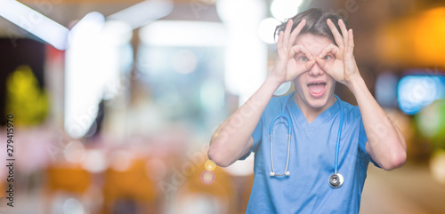Young doctor wearing medical uniform over isolated background doing ok gesture like binoculars sticking tongue out, eyes looking through fingers. Crazy expression.