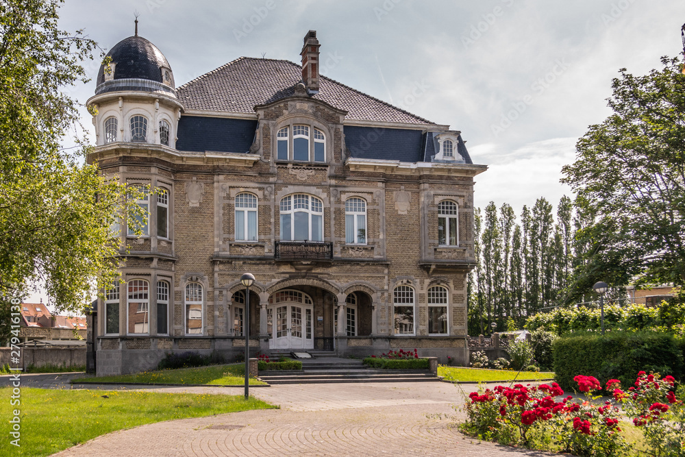 Knokke-Heist, Flanders, Belgium -  June 18, 2019: Kursaalstraat. Historic brown-gray building of Provincial Fishermen School, now defunct, in its park under cloudscape. Green foliage and red flowers.
