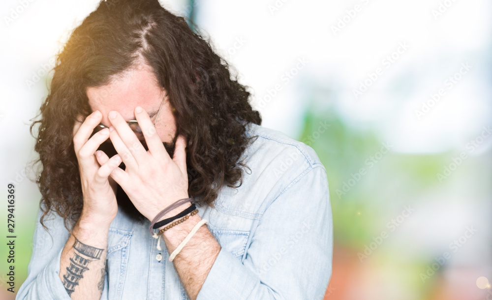 Young hipster man with long hair and beard wearing glasses with sad expression covering face with hands while crying. Depression concept. Young hipster man with long hair and beard wearing glasses with sad expression covering face with hands while crying. Depression concept.