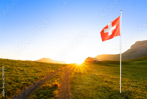 swiss chalet or farm in mountain landscape at sunrise with sun star and swiss flag waving in the foreground 