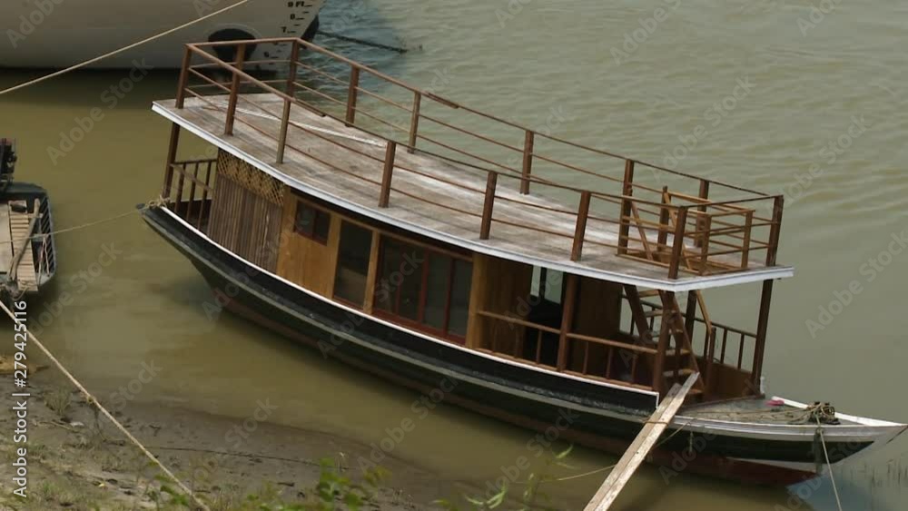 A close up shot of a wooden boat with a shade, stairs and a balcony on the bank of a river A close up shot of a wooden boat with a shade, stairs and a balcony on the bank of a river