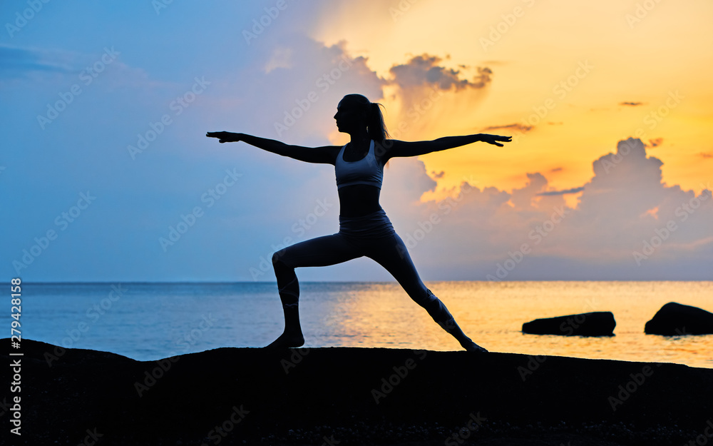 Silhouette of a young woman doing yoga exercises by the sea against the backdrop of colourful dawn	