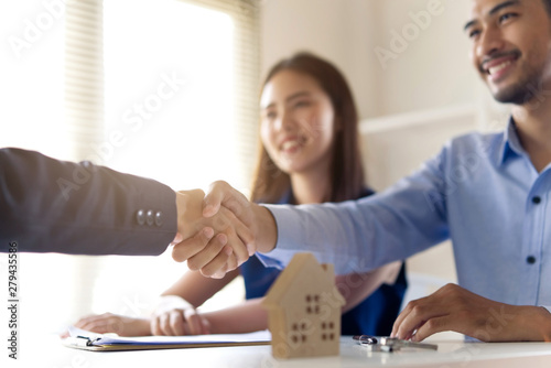 Young Asian couple making contract with house sale agency. Man shaking hand with female agent sitting opposite and his wife sitting next to him with smile looking at house broker. Real estate concept.