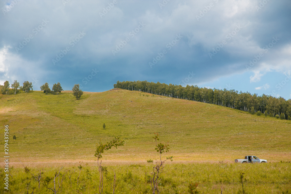 Panorama of nature. The field goes the truck on background of the cloudy sky and the green mountains. There are trees on the mountain.