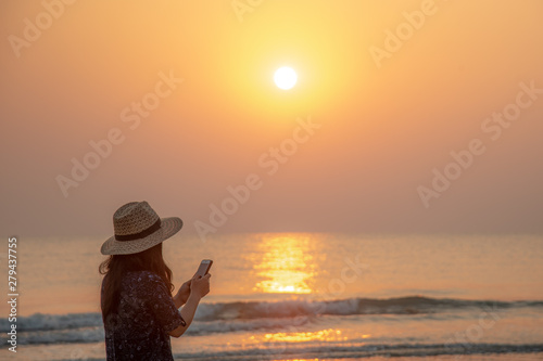 Female traveler using in mobile phone to connect her friend for holiday on sunset background beach seascape thailand.