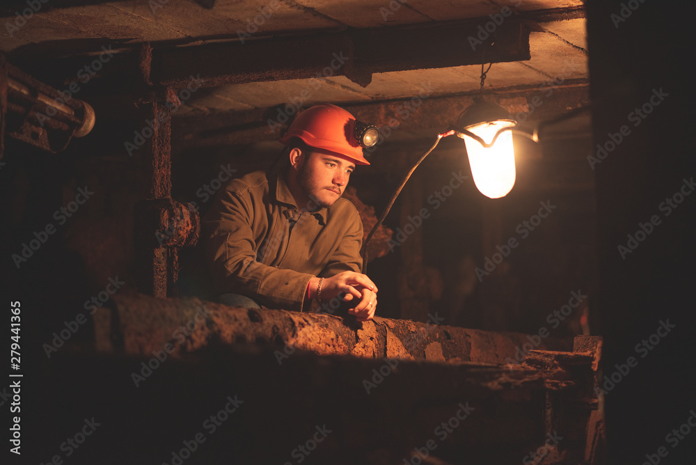 Fototapeta premium A young guy in a protective suit and helmet sits in a tunnel with a burning scrapbook. Miner in mine