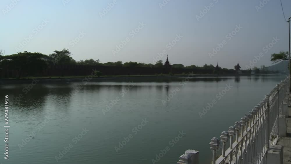 An establishing shot of a walkway close to a pond which passes across a fenced building in the city of Mandalay, Myanmar