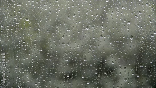 weakly rain water drops fall, hit and flow on glass window, bad windy weather and green trees blur background, cold summer evening, selective focus of closeup full hd stock video footage in real time