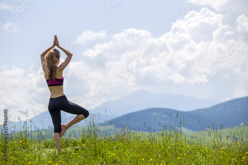 Fototapeta Naklejka Na Ścianę i Meble -  Attractive slim young woman doing yoga exercises outdoors on background of green mountains on sunny summer day.