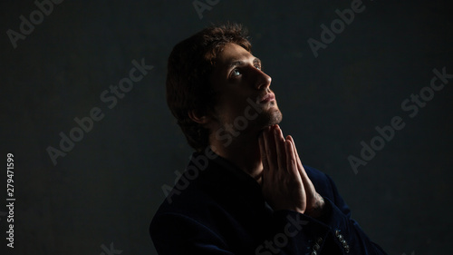 Portrait of a young pensive man on a dark background, looking to the side. Hands in prayer gesture.