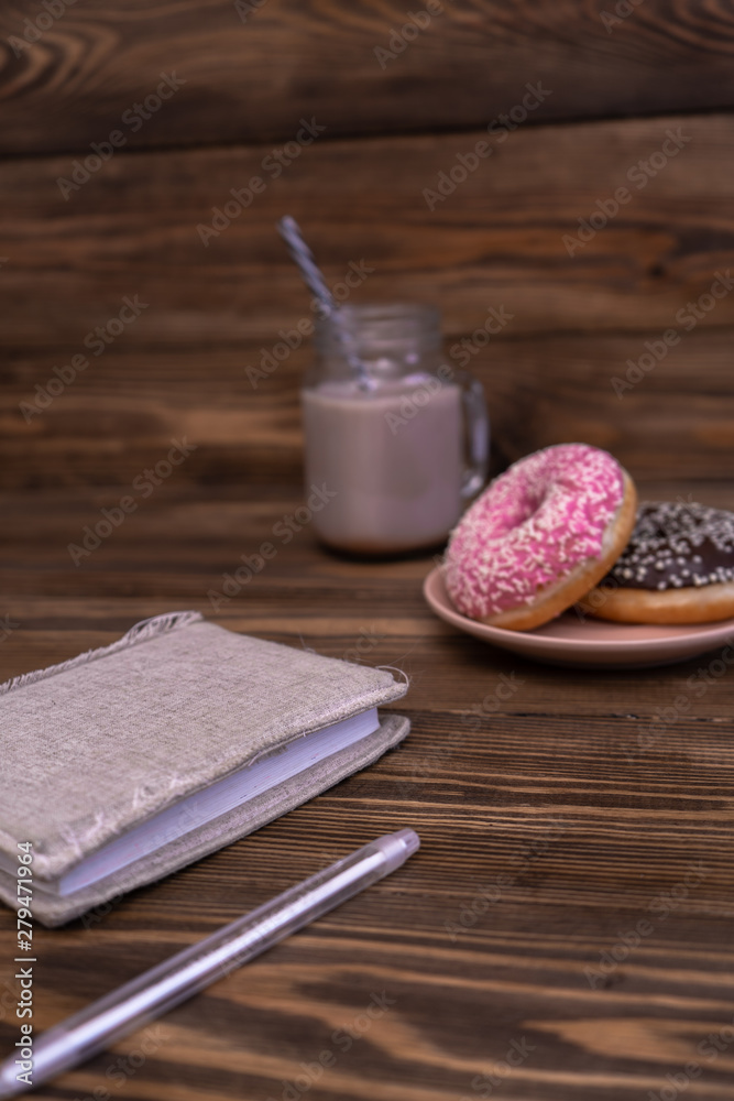 A notebook and a pen, a plate with donuts and a glass mug of coffee on a wooden background. Coffee break. Vertical. Side view A notebook and a pen, a plate with donuts and a glass mug of coffee on a wooden background. Coffee break. Vertical. Side view