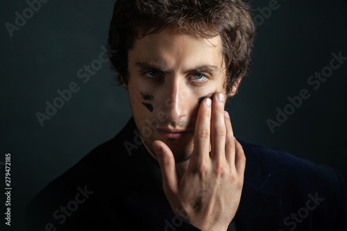 A young handsome man with a slight unshaven face and decisiveness in his eyes, puts a black war paint on his cheeks.