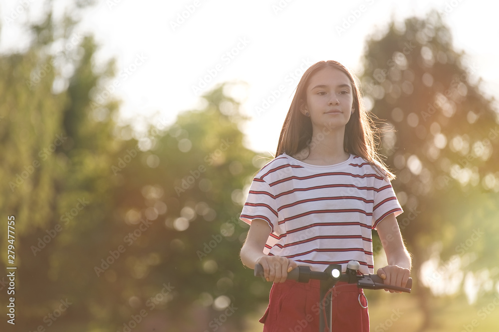 Cute teenager girl riding electric kick scooter in the park at sunset Cute teenager girl riding electric kick scooter in the park at sunset