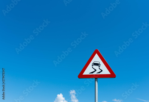 Slippery road traffic sign against blue sky with white clouds, copy space.