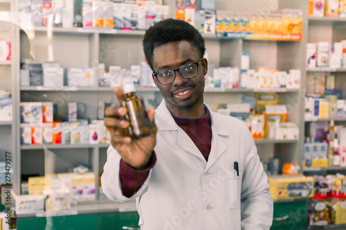 Professional African American male pharmacist showing bottle of medicine according to prescription of customer in modern drugstore