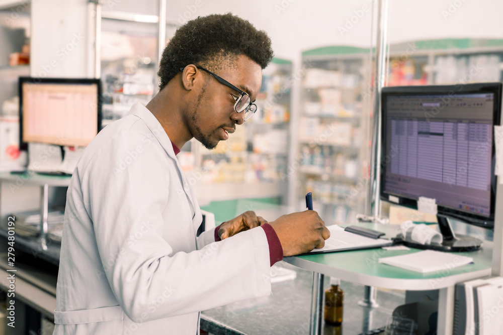 Experienced African American male pharmacist working on pc in modern pharmacy and writing working notes Experienced African American male pharmacist working on pc in modern pharmacy and writing working notes