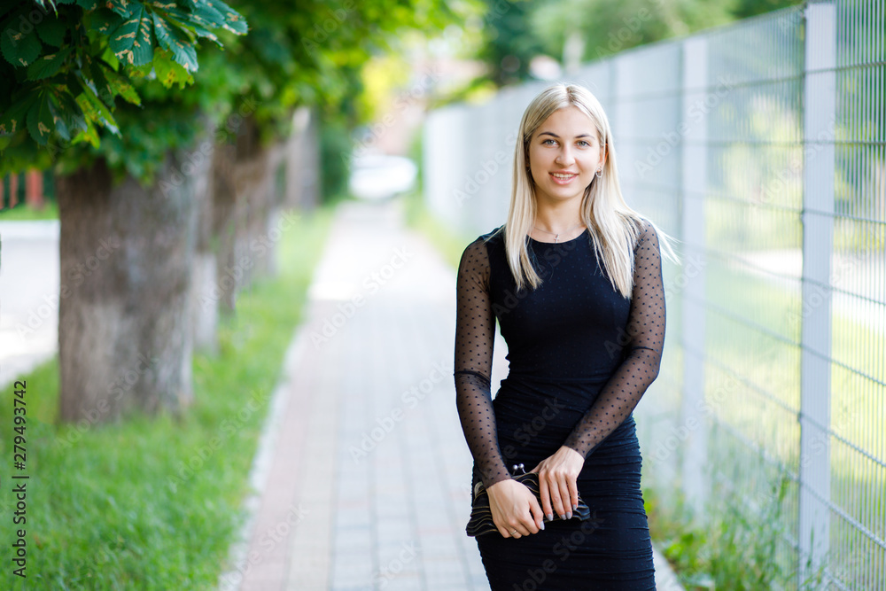The young beautiful blond girl, dressed in a classic black dress, holds a small handbag in her hands, walks through the streets of the city. Business clothing style The young beautiful blond girl, dressed in a classic black dress, holds a small handbag in her hands, walks through the streets of the city. Business clothing style