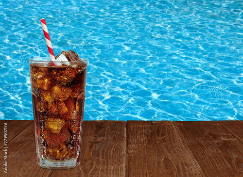 Cola with ice cubes and straw in glass on wooden table with pool background
