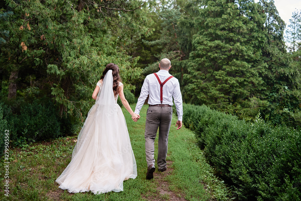 Happy bride and groom holding hands and walking in garden on wedding day. Wedding couple in love, newlyweds, copy space