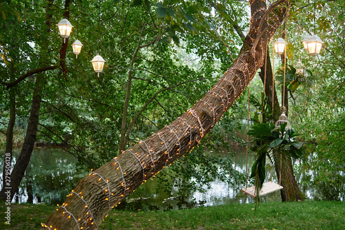 Wedding swing with orchid flowers and monstera palm leaves hanging on tree decorated with lanterns and garland, copy space. Wedding decorations