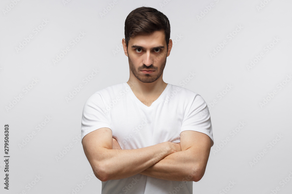Young man in standing with arms crossed and serious concentrated face at camera, looking aggressive, isolated on gray background Young man in standing with arms crossed and serious concentrated face at camera, looking aggressive, isolated on gray background