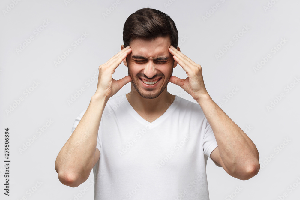 Close-up of young man isolated on gray background in white casual t-shirt touching temples with fingers as if suffering from severe headache Close-up of young man isolated on gray background in white casual t-shirt touching temples with fingers as if suffering from severe headache