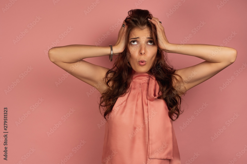 Young brunette holds her head in perplexity with a funny face expression, pink background Young brunette holds her head in perplexity with a funny face expression, pink background