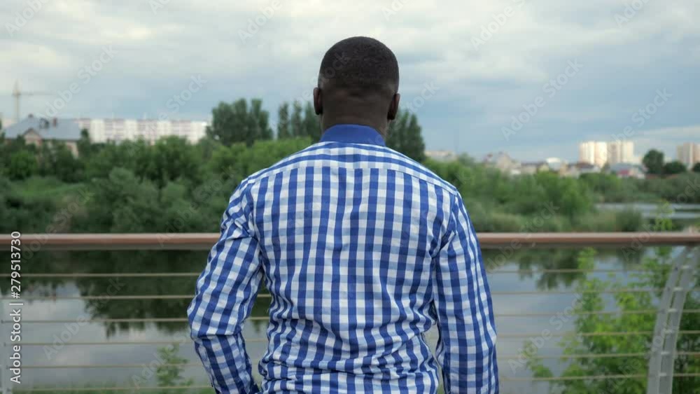 Young afro american guy on walking in park. Black bearded man is coming to waterfront and admiring cityscape, back view. Young afro american guy on walking in park. Black bearded man is coming to waterfront and admiring cityscape, back view.