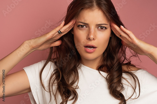 Close-up of a young brunette holding her head with tension over pink background