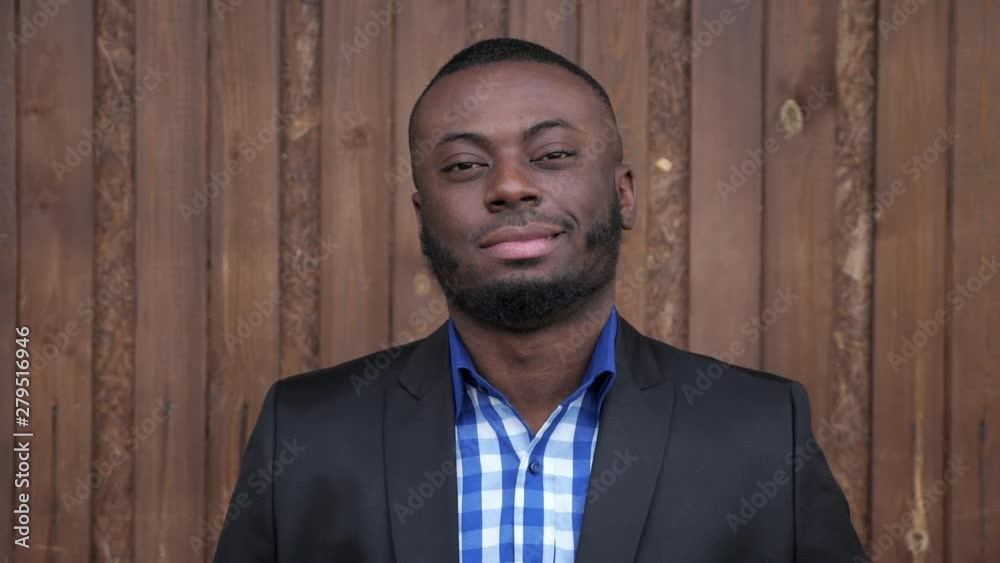 Portrait of young black bearded guy. Afro american man in suit is smiling and looking at camera on dark wood background. He is happy and confident.