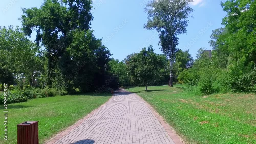 First person view or perspective walking down the foot path in the park with green trees grass and bushes on sunny summer day