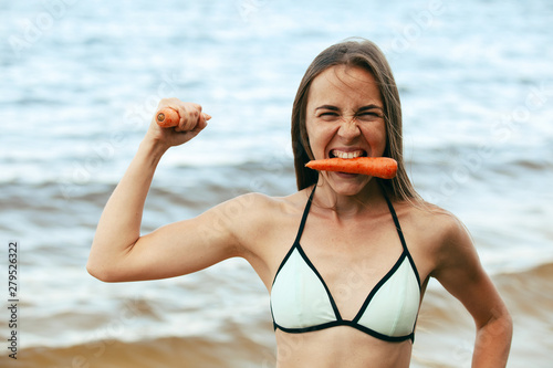 Happy vegan girl with a carrot in the mouth and hand showing a power of a healthy food.