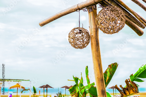 Exotic view of the sea horizon, sandy beach, beach umbrellas and sun loungers from the sea cafe with a wicker lamp, under a wooden rustic-style roof, banana tree leaves on a sunny day in the tropics