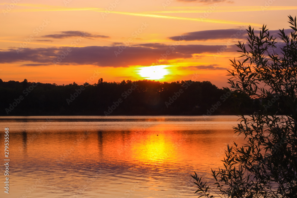 Fototapeta premium Beautiful landscape with orange sunset over the water. Vegetations in foreground and symmetry of the colorful clouds in a lake. 