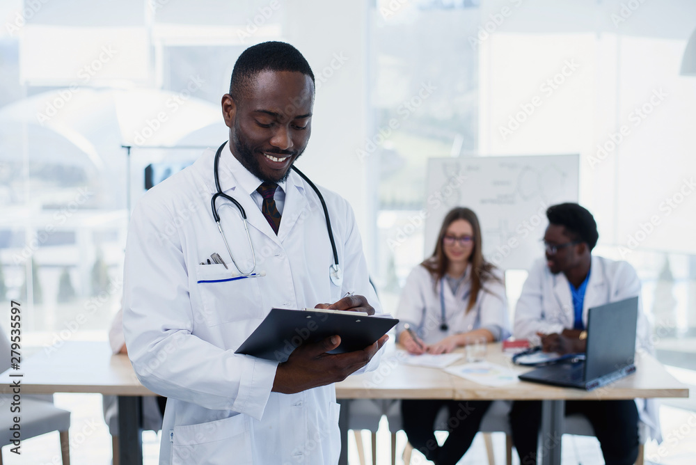 Handsome Afro American doctor in white coat makes some notes. Young medical student with a stethoscope around his neck holds a folder. Handsome Afro American doctor in white coat makes some notes. Young medical student with a stethoscope around his neck holds a folder.