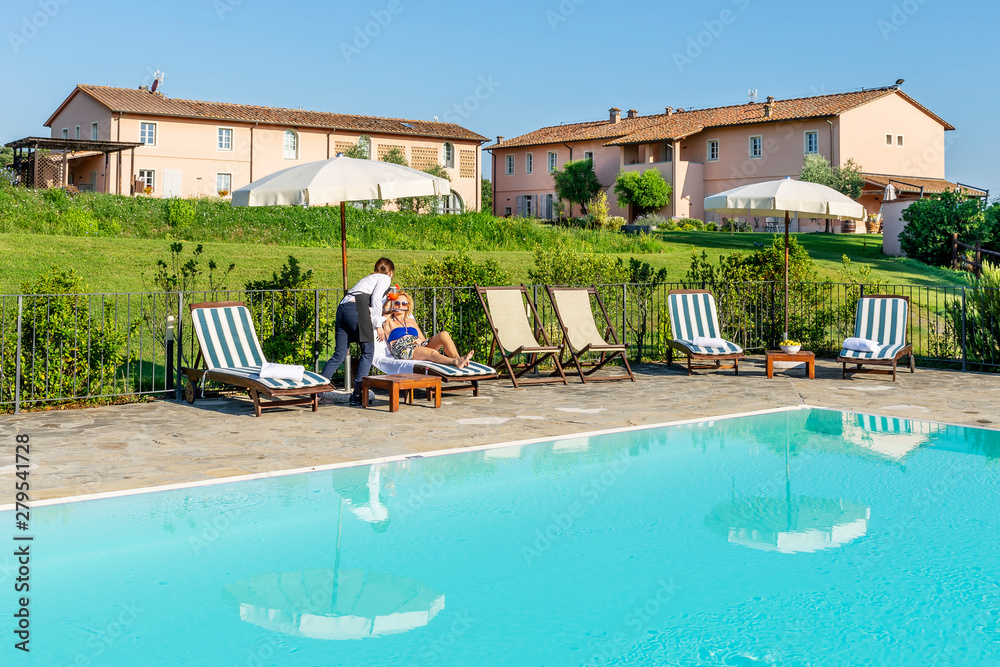 Young waitress serves a poolside cocktail to a customer sitting on a lounger in a resort in the countryside of Pisa, Tuscany, Italy