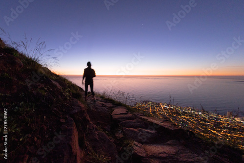 The silhouette of a hiker walking off a mountain at night above a city and its lights