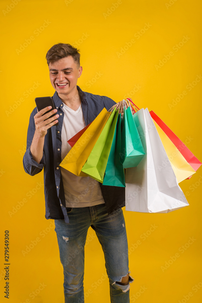 Handsome guy with shopping bags is using a mobile phone and smiling. Portrait of a smiling man holding shopping bag over yellow background and looking at camera Handsome guy with shopping bags is using a mobile phone and smiling. Portrait of a smiling man holding shopping bag over yellow background and looking at camera