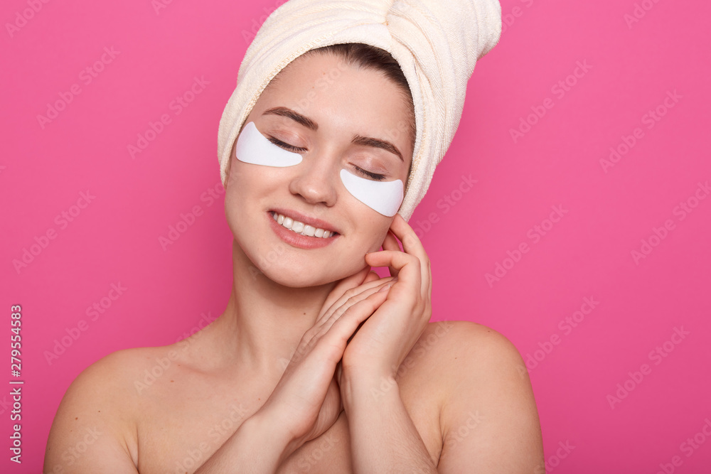 Image of cheerful peaceful young woman having pleasant facial expression, smiling sincerely, posing with closed eyes, touching her face with hands, being naked, having patches on face. Beauty concept. Image of cheerful peaceful young woman having pleasant facial expression, smiling sincerely, posing with closed eyes, touching her face with hands, being naked, having patches on face. Beauty concept.
