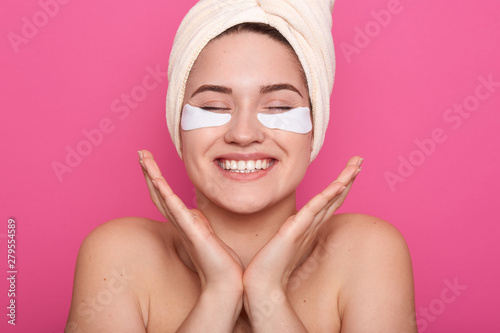 Indoor picture of positive energetic European woman keeping hands together near face, closing eyes, laughing sincerely, being fond of beauty procedures, having patches under eyes, waiting for results.
