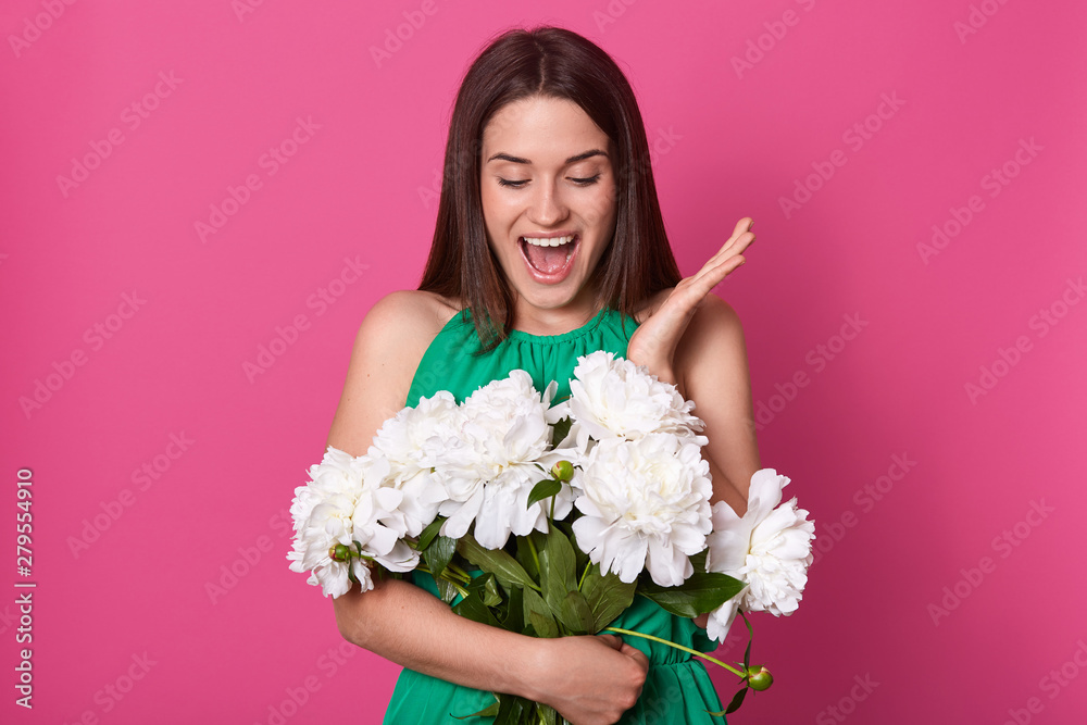 Portrait of cheerful happy young woman holding bouquet of white peonies, looking at them with surprise, raising one hand up, opening her mouth widely, being in high spirits, getting present.