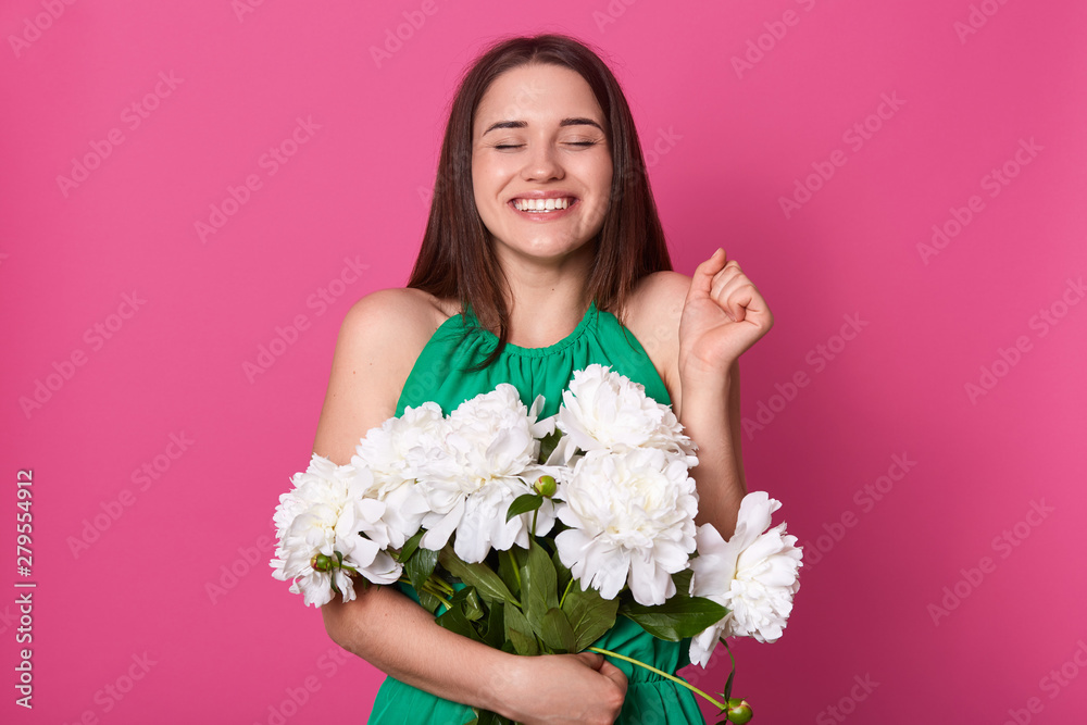 Image of good looking positive model posing isolated over pink background in studio, closing her eyes, raising one hand, holding white peonies, smiling sincerely, wearing green summer dress.