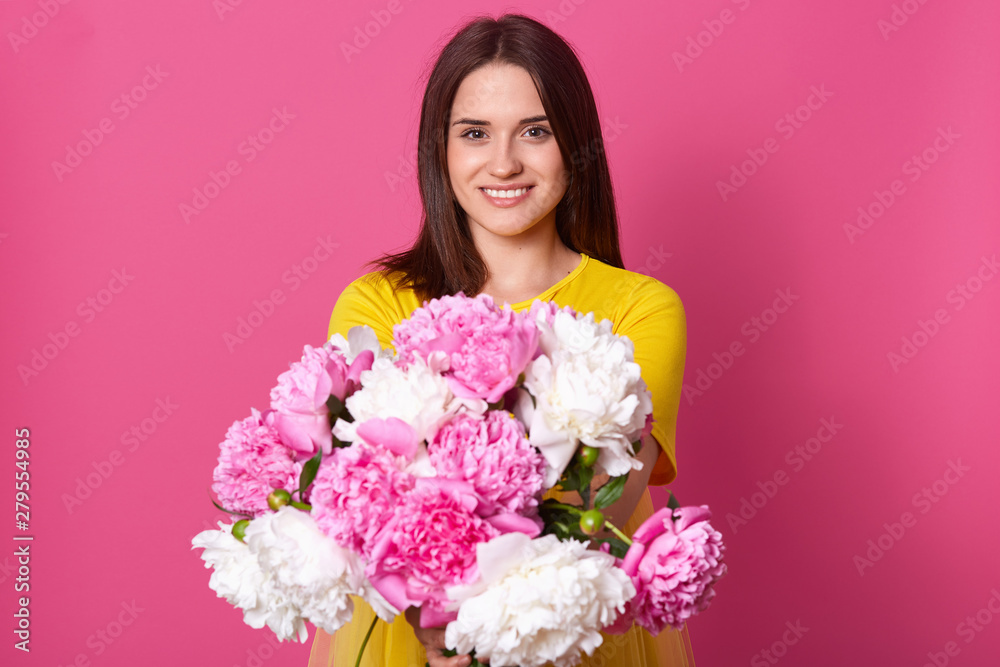 Image of cheerful amazing model smiling sincerely, looking directly at camera, holding peonies in distance, getting flowers for present, wearing bright yellow dress. People and emotions concept.