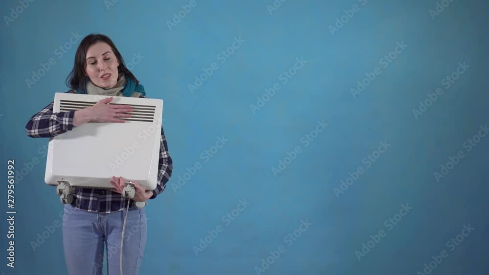 Frozen woman in scarf holding in his hand an electric heater on a blue background copy space Frozen woman in scarf holding in his hand an electric heater on a blue background copy space