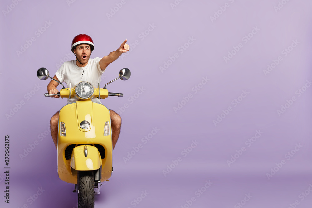Scared man drives fast yellow scooter, wears protective helmet and white t  shirt, points index finger in distance, notices scarying scene on road,  poses against purple wall, blank copy space. Driving Stock