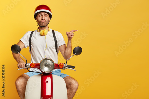 Fototapeta Funny unshaven guy in casual wear, drives on motorcycle, has good trip, enjoys freedom, points index finger on blank space over yellow wall, pouts lips