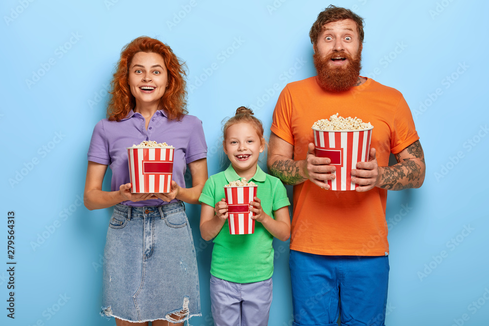 Loving ginger family of three members eat popcorn while watch TV programme, look happily at camera. Mother, father and daughter spend leisure time at local cinema, enjoy watching funny comedy Loving ginger family of three members eat popcorn while watch TV programme, look happily at camera. Mother, father and daughter spend leisure time at local cinema, enjoy watching funny comedy