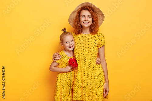 Love you mum! Pretty ginger girl with red flower, embraces mother, enjoy good time together. Fashionable two sisters in same dresses pose over studio yellow wall. Motherhood, parenthood concept