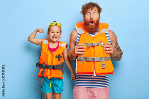 Father and daughter in safety lifejackets going to have extreme activity on water. Shocked bearded ginger man stares at camera, cheerful little girl shows biceps, have summer rest together. Family