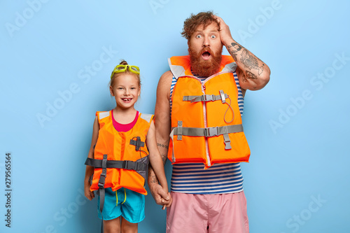 Horizontal shot of amazed bearded redhead father in orange inflated lifevest, holds hand of small child, have sea cruise during summer, stand together against blue background. Friendly family